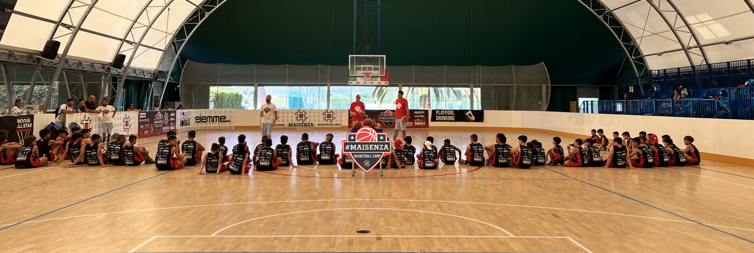 Panoramica dei ragazzi del camp seduti in fila sul parquet sotto la tensostruttura con il cartello #MAISENZA