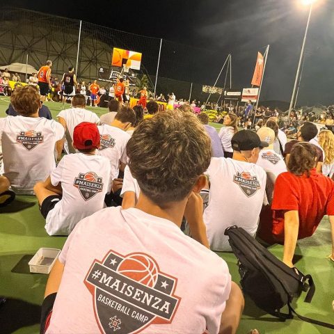 Ragazzi del Maisenza Basketball Camp di spalle con la t-shirt #MAISENZA, seduti a bordo campo durante una serata di basket